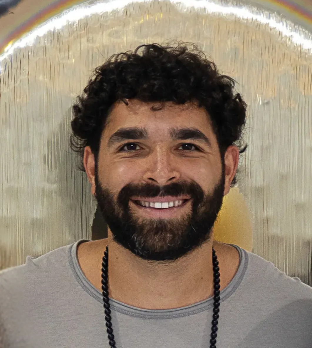 Smiling man with dark curly hair and beard, wearing a gray shirt and black beaded necklace, in front of a reflective backdrop.