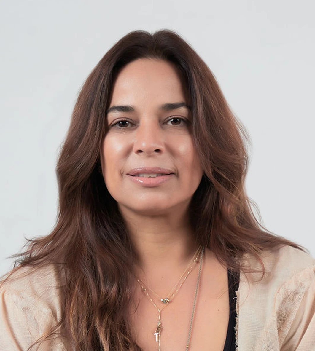 Smiling woman with long brown hair, wearing a beige cardigan and layered necklaces against a light gray background.
