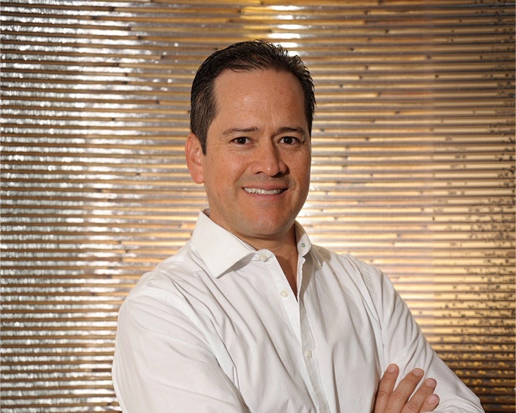 Portrait of a smiling man in a white button-down shirt with arms crossed, standing against a metallic striped background on a reflective wall.