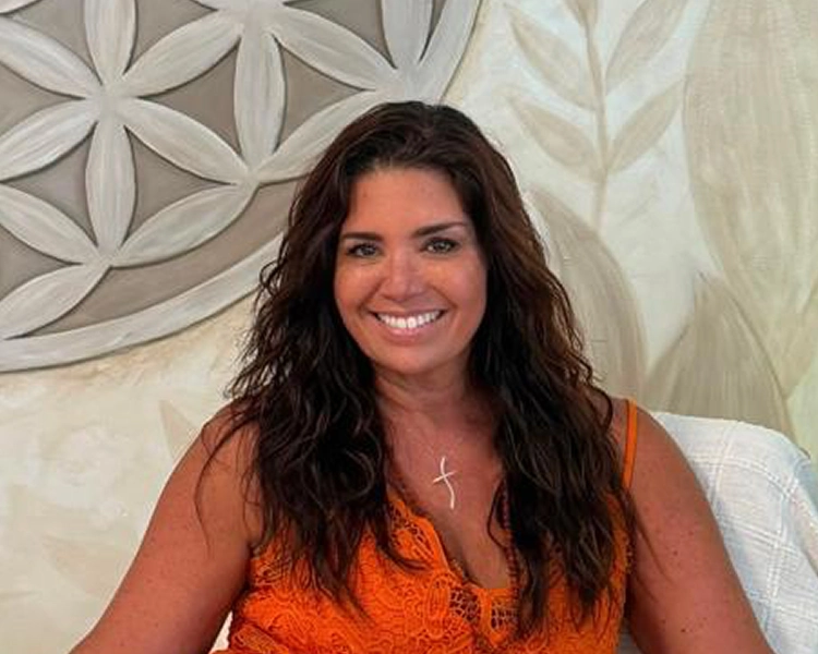 Smiling woman with long wavy brown hair wearing an orange lace top and a cross necklace, seated indoors against a light patterned wall background.